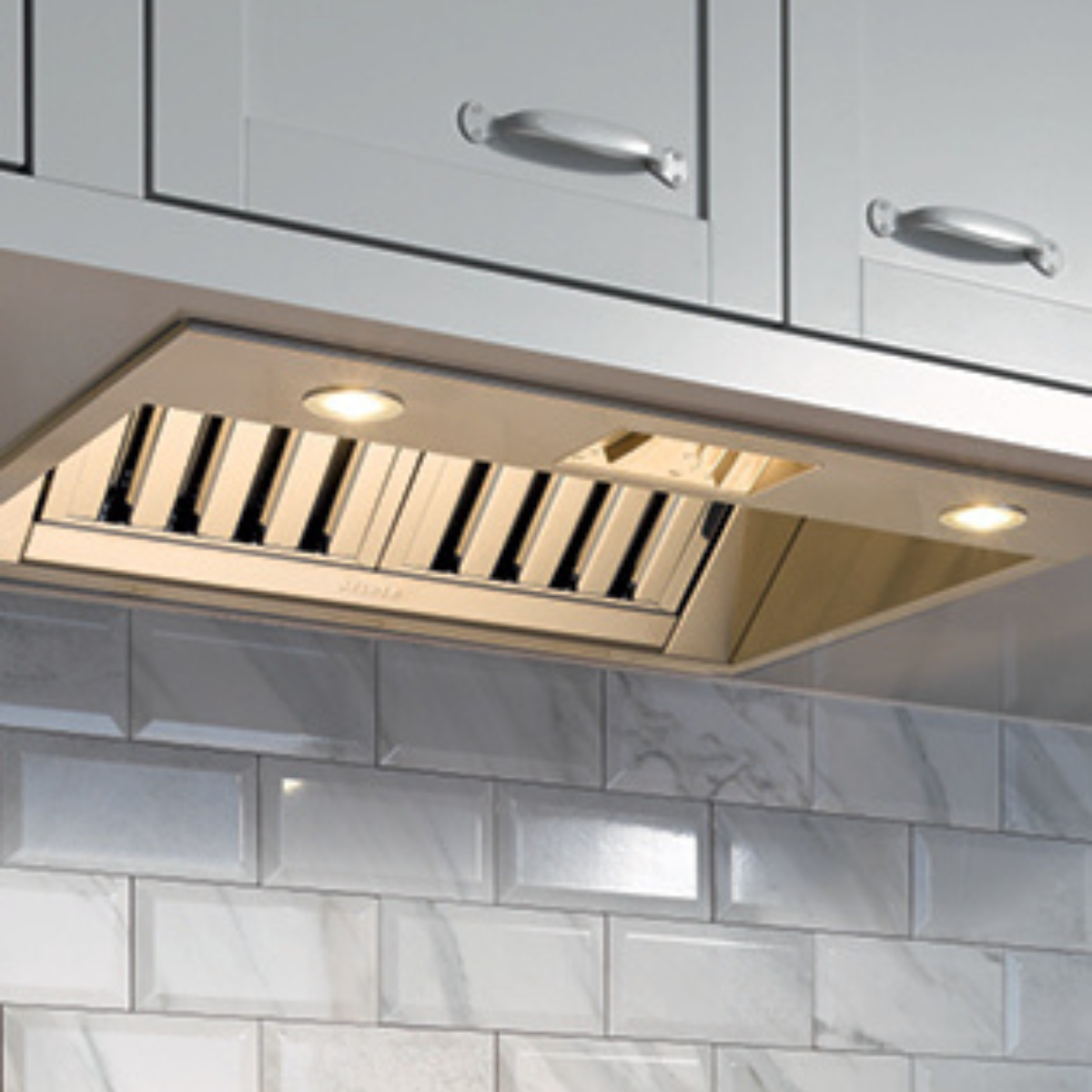 Close-up of a brass-toned under-cabinet range hood insert with baffle filters, installed above a marble tile backsplash.