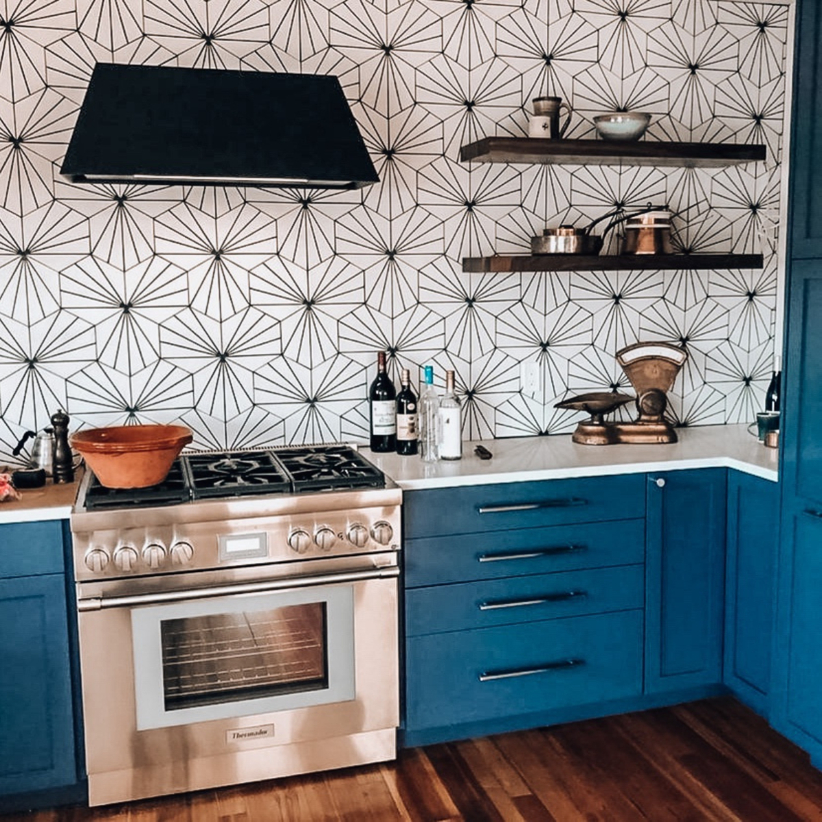 Blue kitchen cabinetry with a black angled range hood above a stainless steel gas range, set against geometric patterned wallpaper.