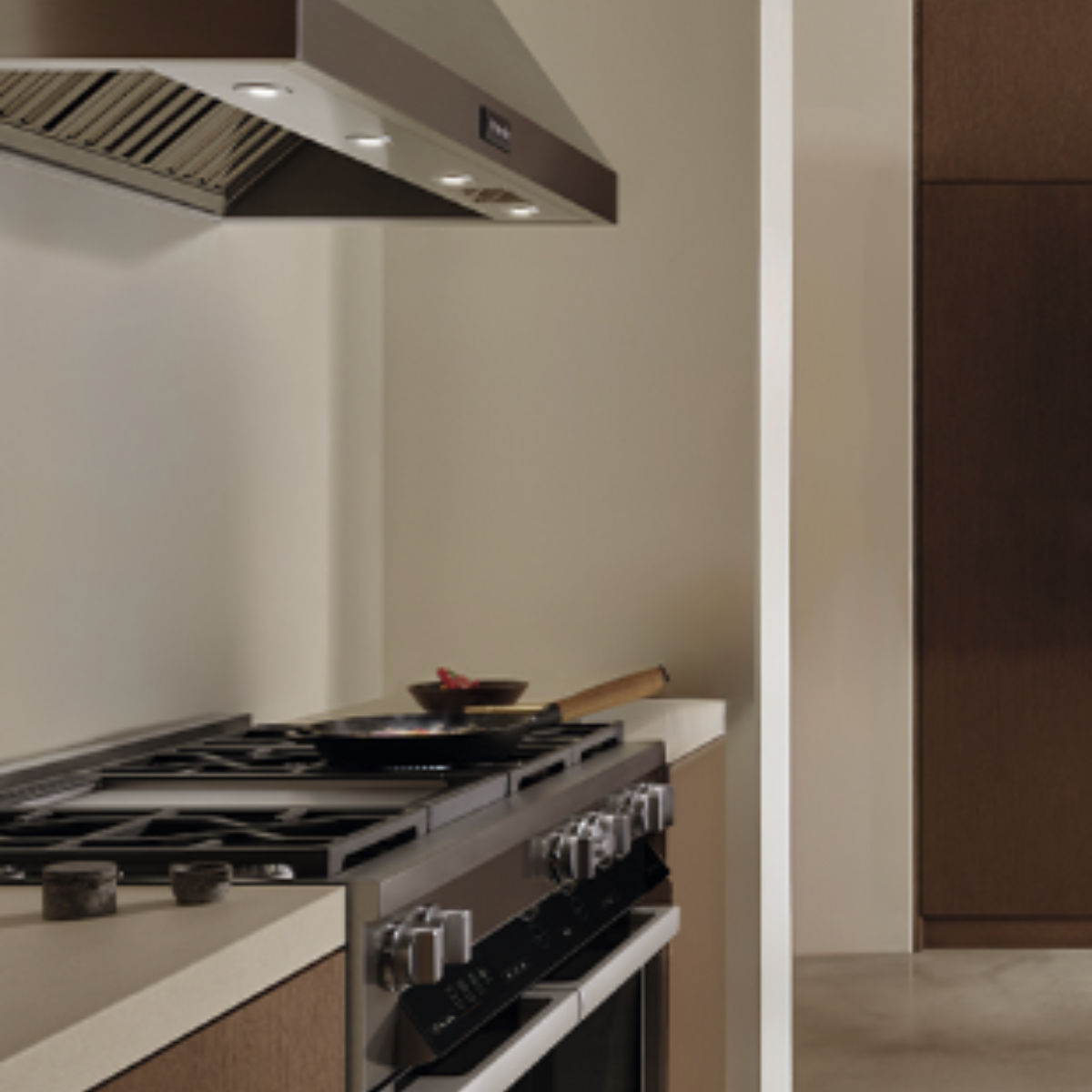 Wall-mounted range hood above a gas cooktop with a pan of vegetables, in a dark wood and neutral-toned kitchen.