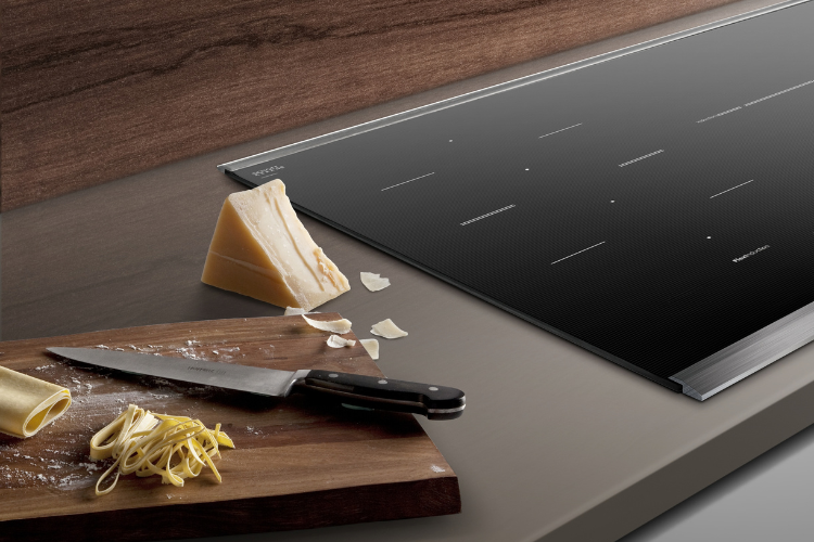 Black induction cooktop set into a taupe countertop beside a wooden board with fresh pasta, a knife, and shaved Parmesan