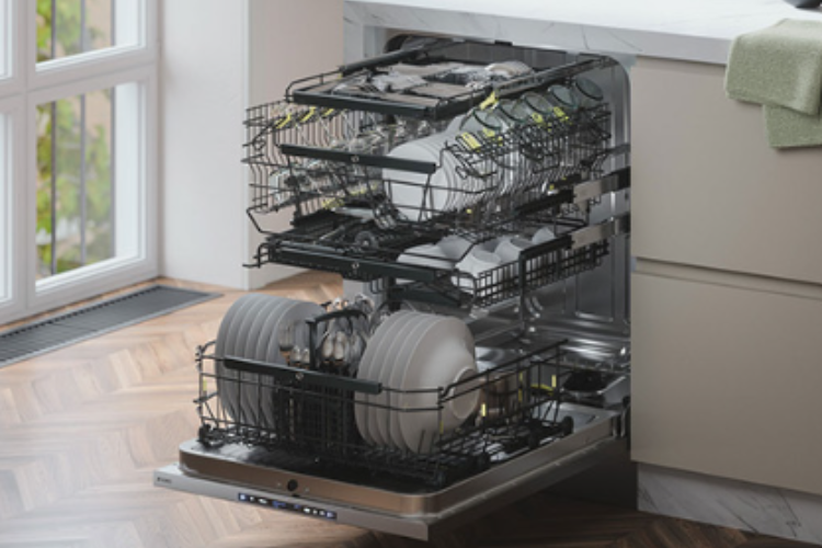 Open dishwasher with three pull-out racks, the top tray holding cutlery above loaded plates, set against a herringbone wood floor.