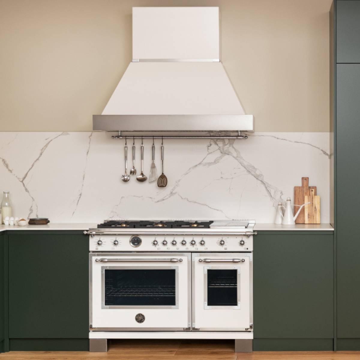  A Bertazzoni dual-oven range in white with six gas burners, chrome hardware, and a matching wall-mount hood, set against a marble backsplash in a kitchen with deep forest green cabinetry.