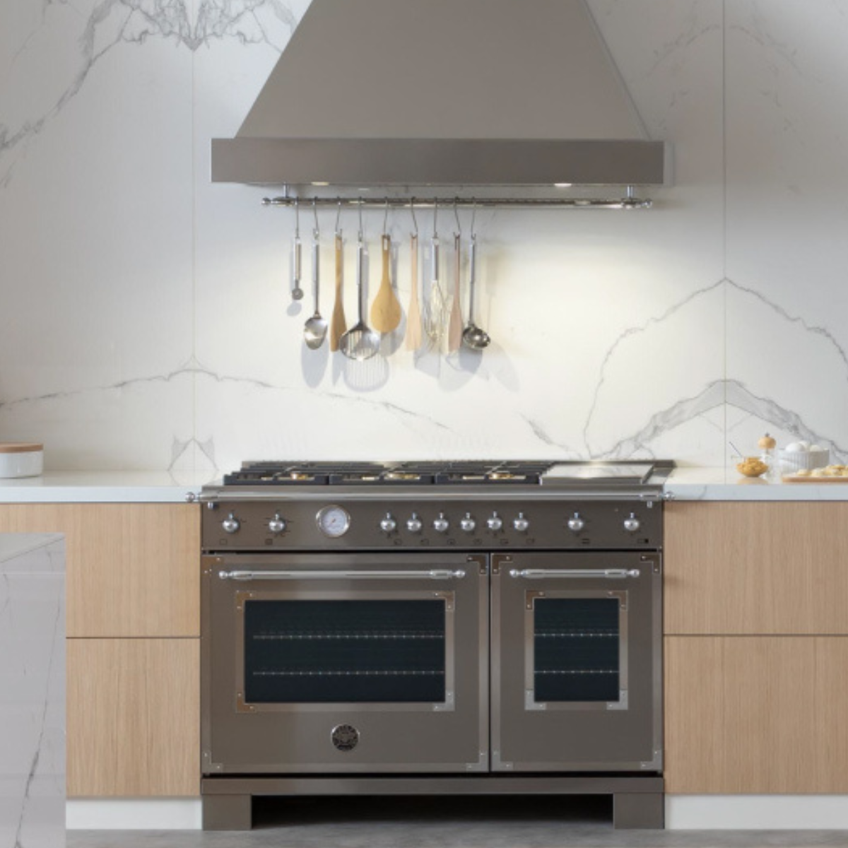 A Bertazzoni dual-oven range in matte brown with six gas burners and a griddle, set into a modern kitchen with light wood cabinetry, white marble backsplash, and a matching wall-mount hood with hanging utensils.