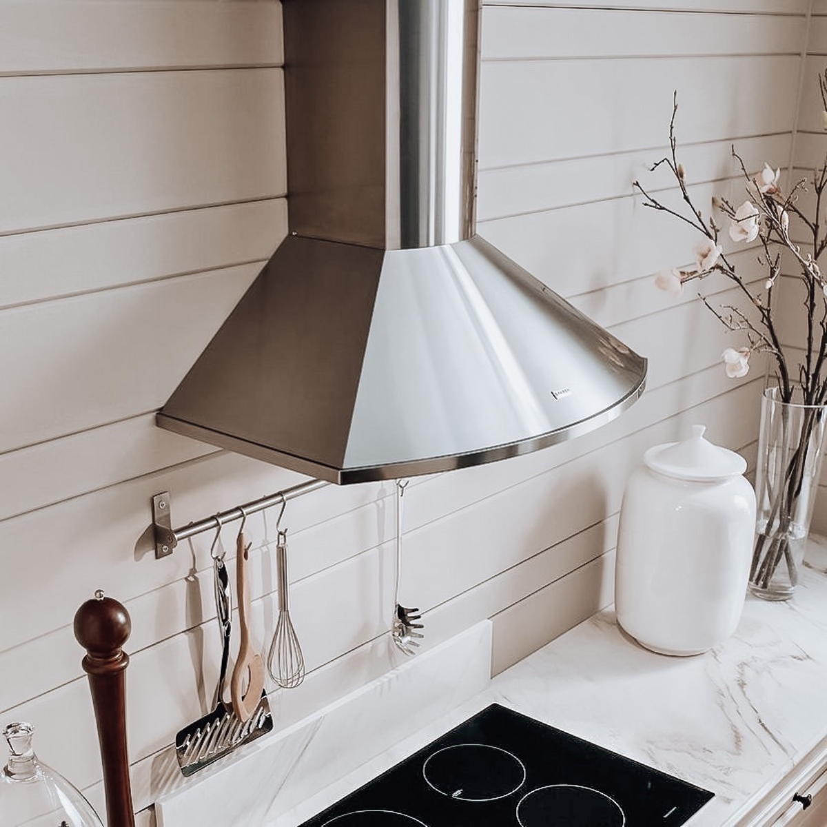A stainless steel wall-mount range hood with a classic pyramid silhouette installed above a black ceramic cooktop in a bright white kitchen with shiplap walls, marble countertops, and hanging utensils on a rail below the hood.