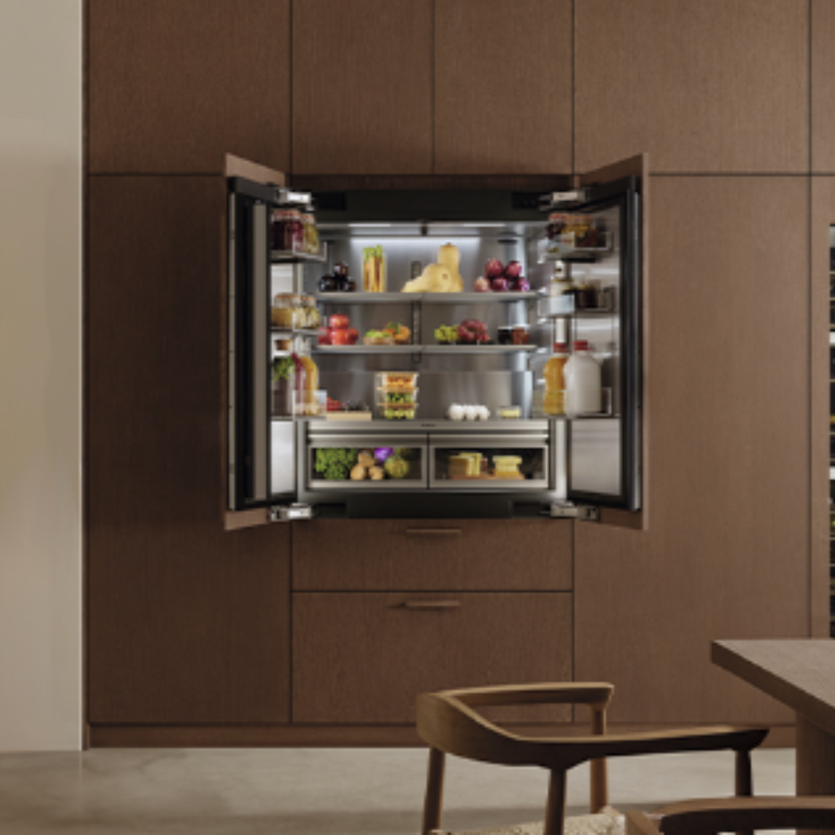  A fully stocked panel-ready French door refrigerator open to reveal illuminated interior shelving, produce drawers, and tall door storage, seamlessly integrated into floor-to-ceiling walnut cabinetry.