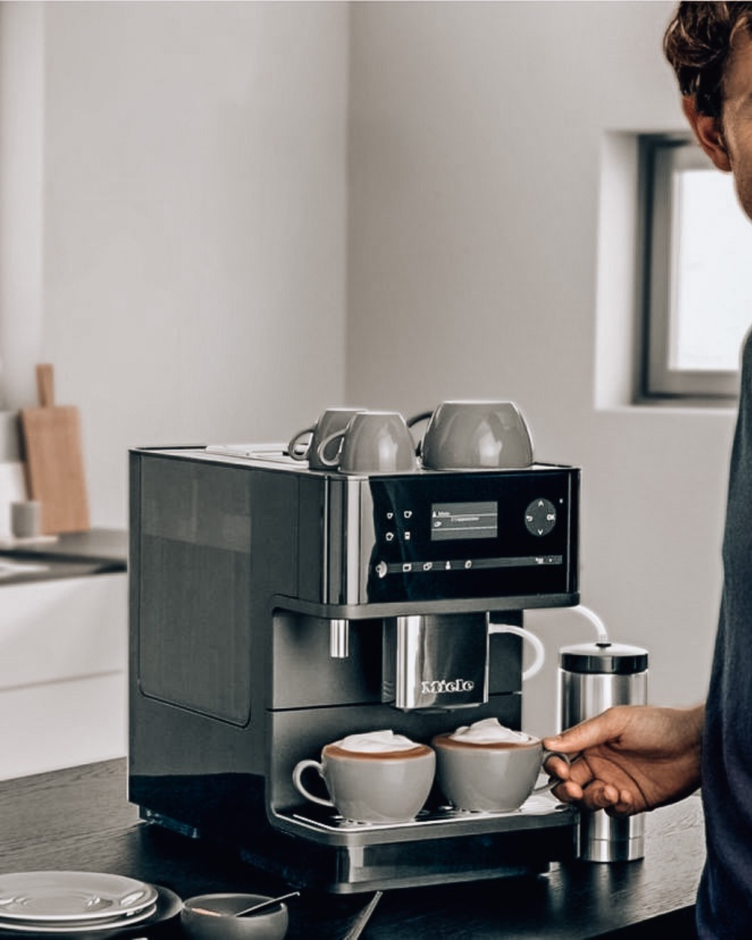 Miele built-in coffee machine dispensing two lattes with milk foam while a person reaches for a finished cup in a bright kitchen.
