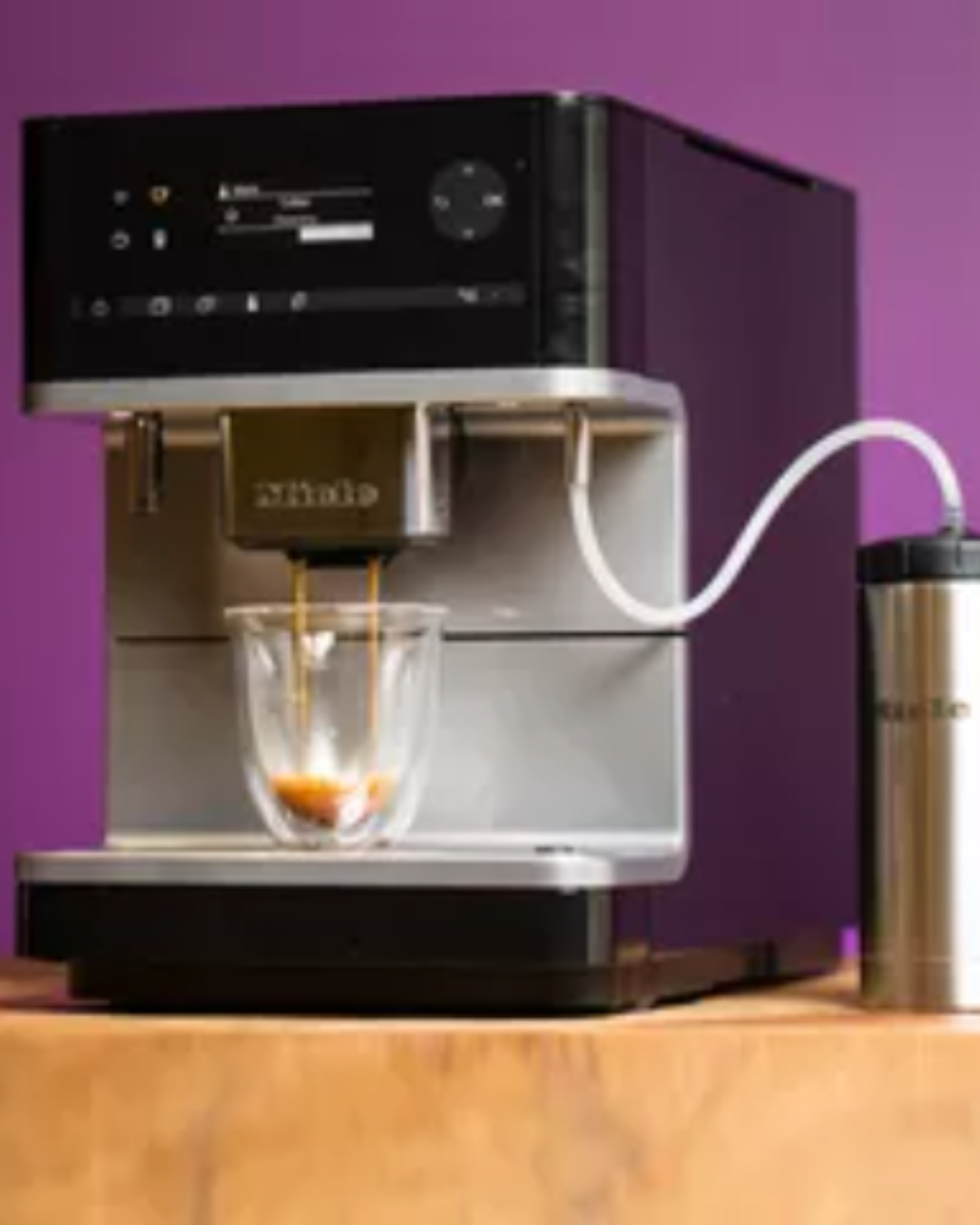 Miele built-in coffee machine brewing espresso into a glass cup, paired with a stainless travel mug, against a purple backdrop.