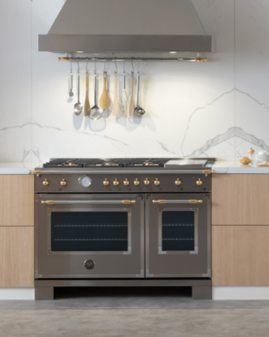 Dark stainless Bertazzoni double-oven range with brass hardware, gas cooktop with griddle, and matching range hood with hanging utensil rail.